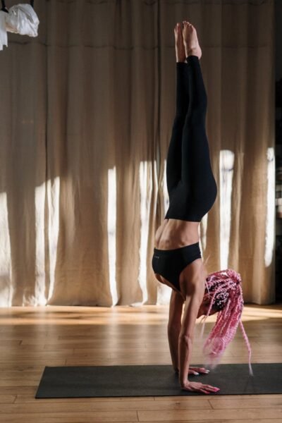 A woman with pink hair practices a handstand on a yoga mat in a serene studio setting.